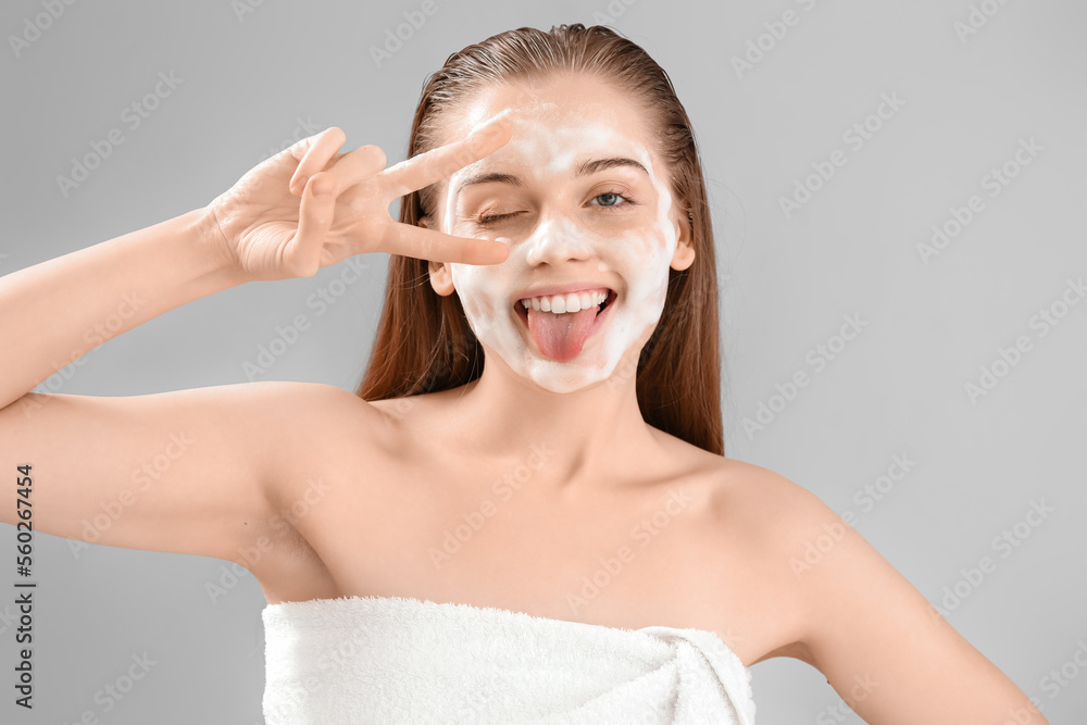 Funny young woman with soap foam on face against grey background