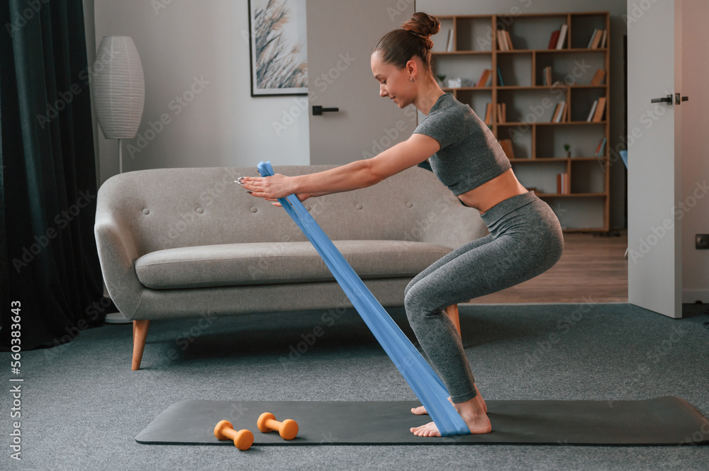 Squats and using blue cloth. Young woman is doing fitness indoors