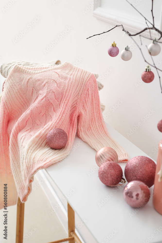 Knitted sweaters and Christmas balls on table near light wall