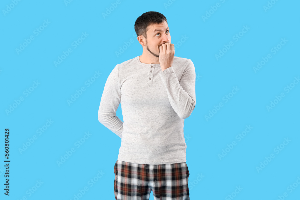 Handsome man in pajamas biting nails on blue background