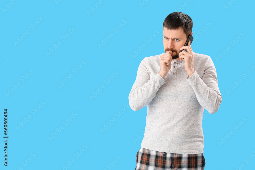 Handsome man biting nails and talking by mobile phone on blue background