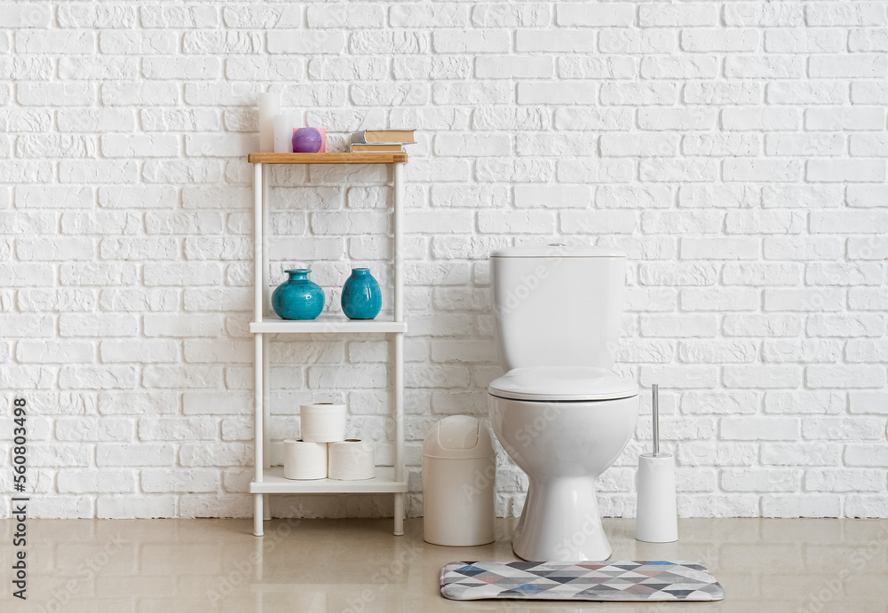 Interior of restroom with ceramic toilet bowl, shelving unit and brush near white brick wall