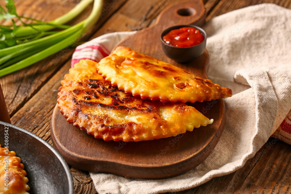 Cutting board with tasty chebureks and sauce on wooden background