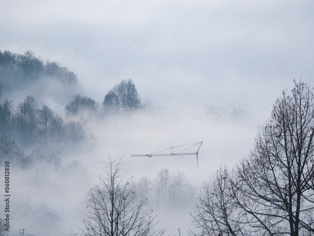 Tall construction crane rising from the winter mist rolling over countryside