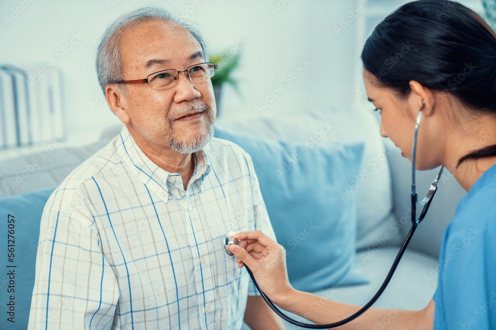 Caring young female doctor examining her contented senior patient with stethoscope in living room. M