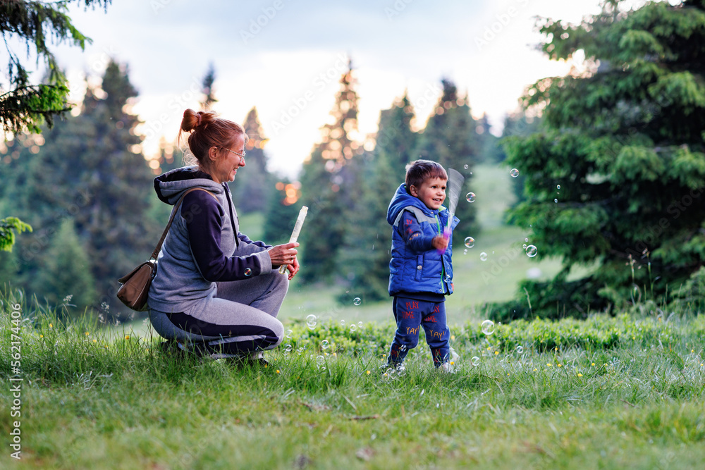 Mother plays with son on a lawn and teaches him to blow soap bubbles against the backdrop of forest