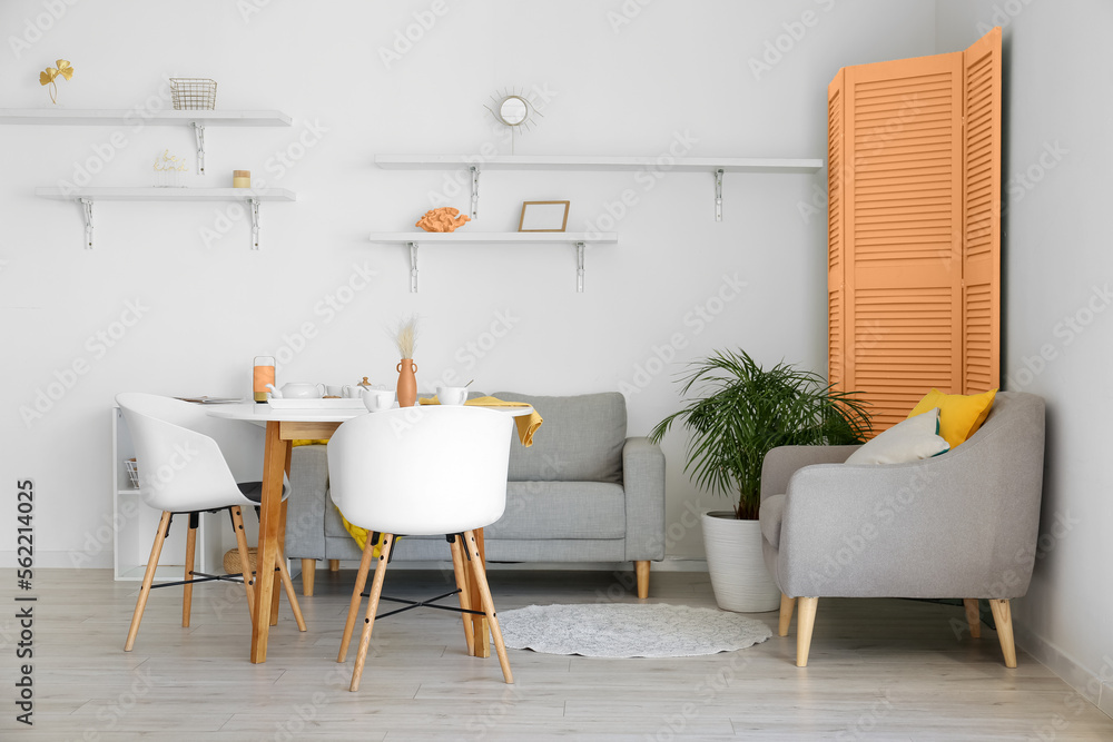 Interior of light dining room with table, grey furniture and orange folding screen