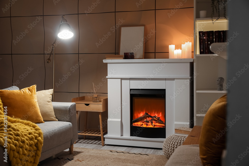 Interior of dark living room with electric fireplace, burning candles and glowing lamp