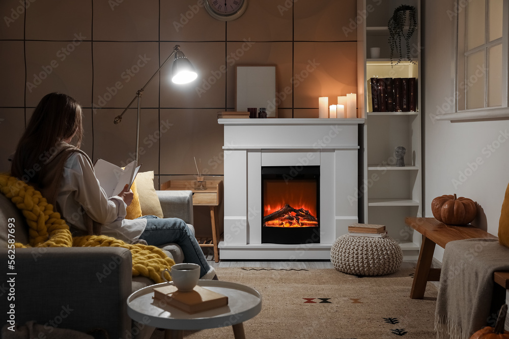 Young woman reading book near fireplace at home in evening