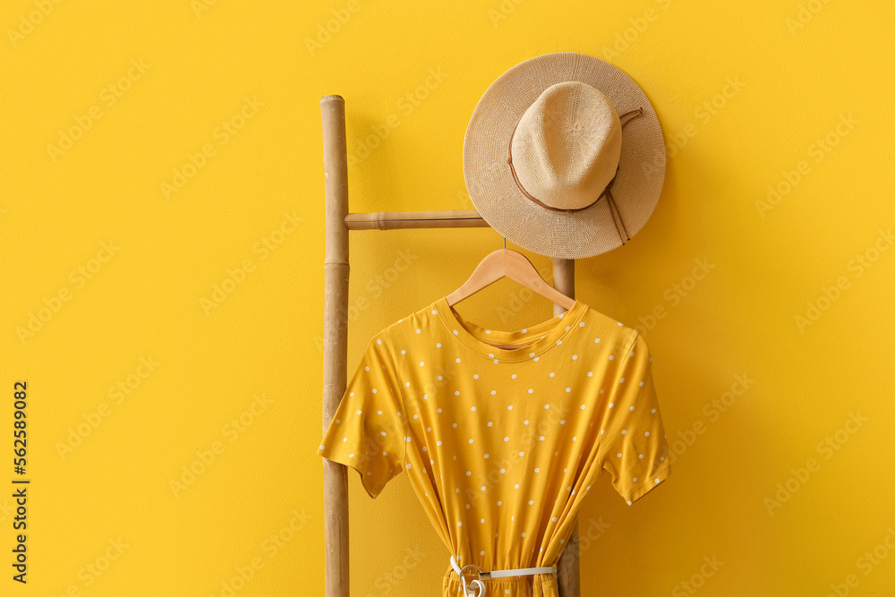 Ladder with stylish clothes and hat near yellow wall