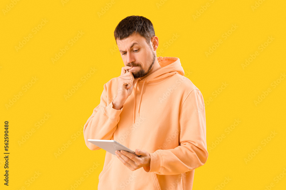 Handsome man with tablet computer biting nails on yellow background