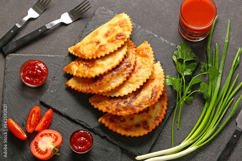 Boards with tasty chebureks, sauces, tomato, onion, juice and forks on dark background