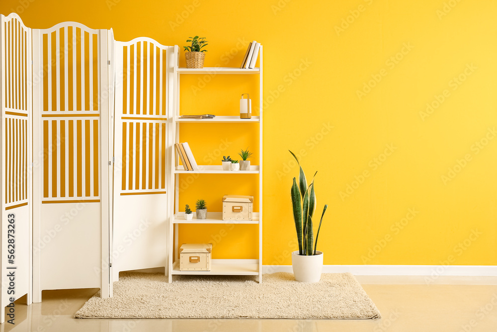 Interior of room with folding screen, shelving unit and houseplant near yellow wall