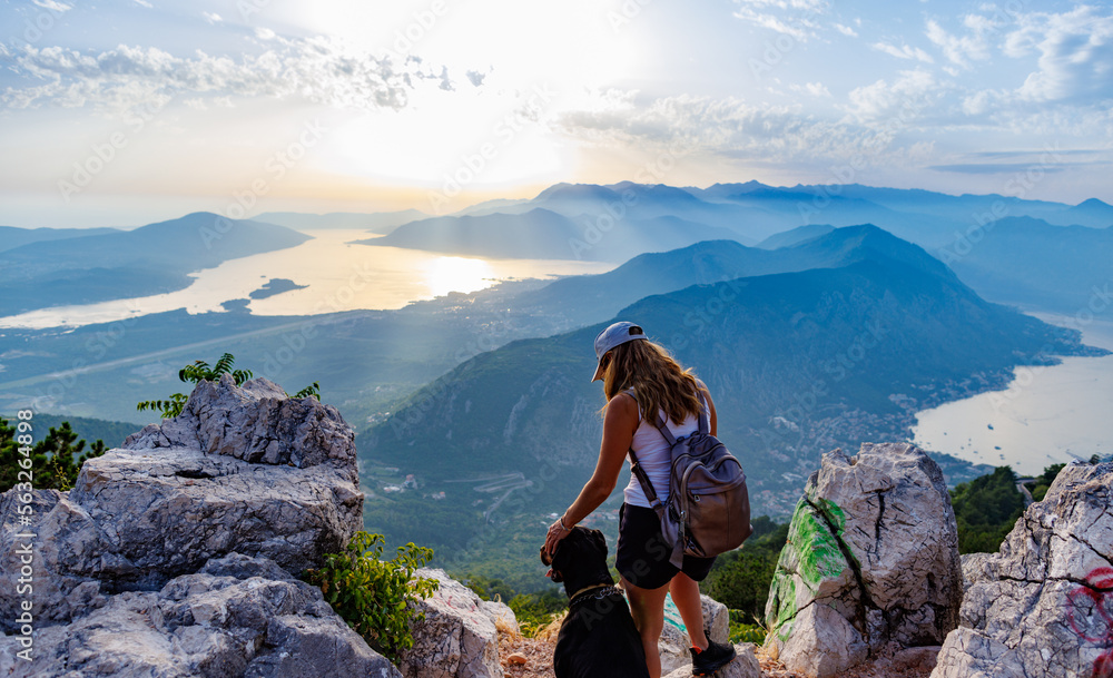 A sporty girl with a backpack stands on the edge of a mountain with a Rottweiler dog