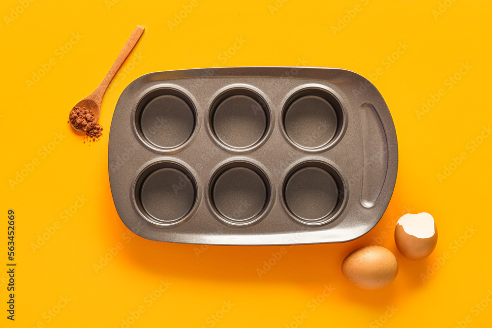 Muffin baking tray with spoon of cocoa powder and eggs on orange background