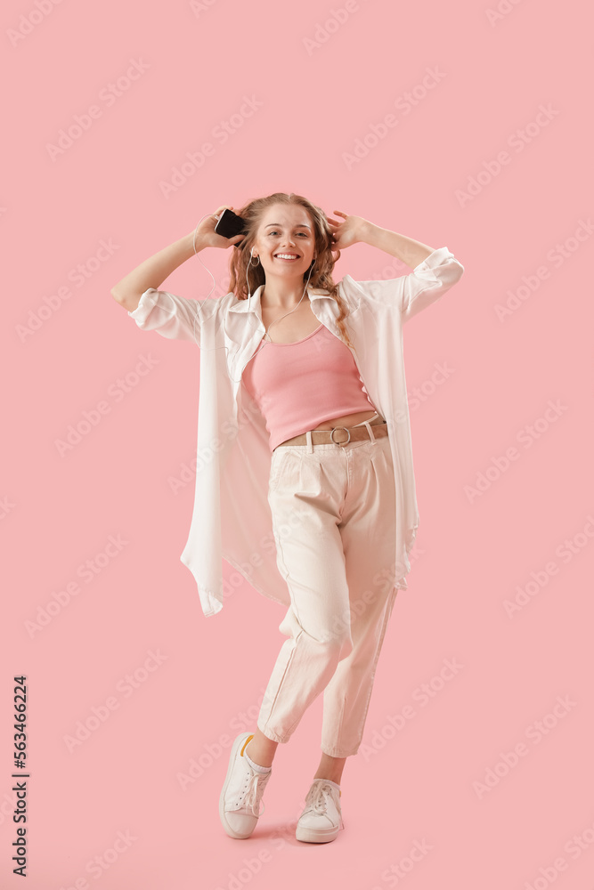 Young woman in earphones with mobile phone dancing on pink background