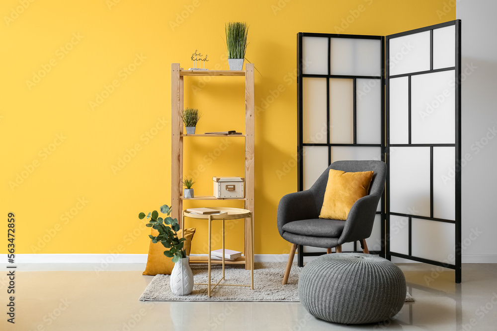 Interior of room with folding screen, armchair and pouf near yellow wall