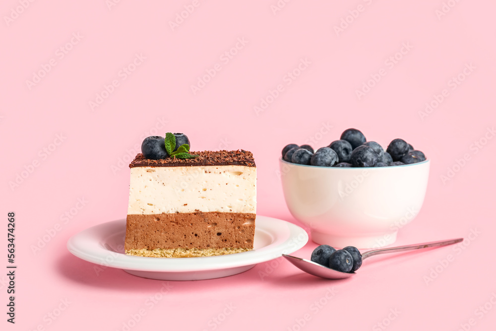 Plate with piece of birds milk cake, bowl of blueberries and spoon on pink background