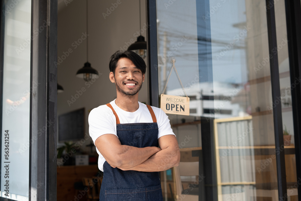 Portrait of smiling owner standing at his restaurant gate with open signboard. Young entrepreneur le