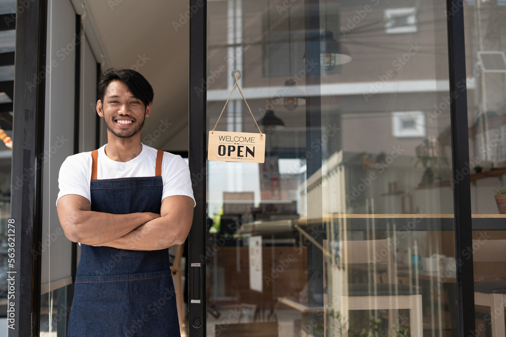 Portrait of smiling owner standing at his restaurant gate with open signboard. Young entrepreneur le