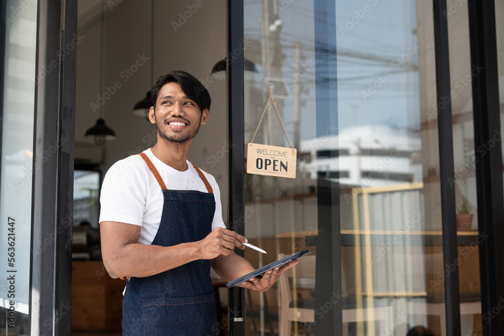 Portrait of smiling owner standing at his restaurant gate with open signboard. Young entrepreneur le