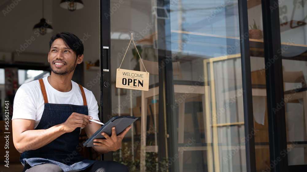 Portrait of smiling owner standing at his restaurant gate with open signboard. Young entrepreneur le