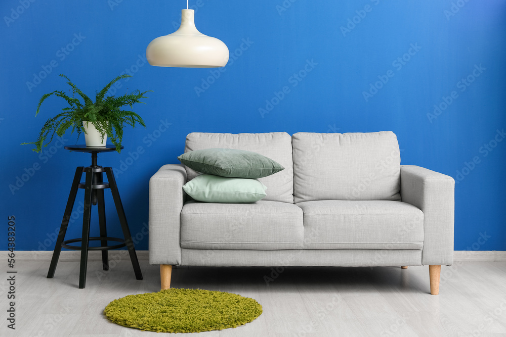 Grey sofa with cushions and houseplant on stool near blue wall