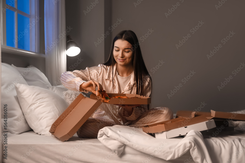 Young woman taking slice of tasty pizza from box in bedroom at night