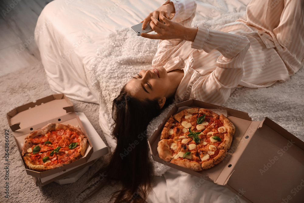 Young woman with tasty pizza using mobile phone in bedroom at night