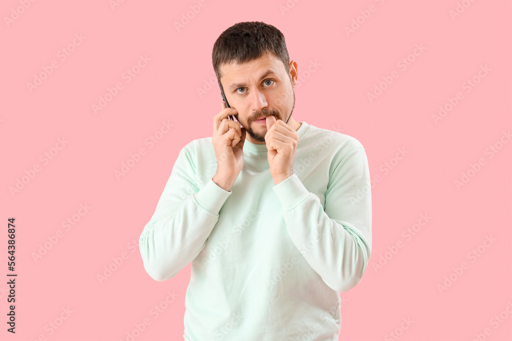 Handsome man biting nails and talking by mobile phone on pink background