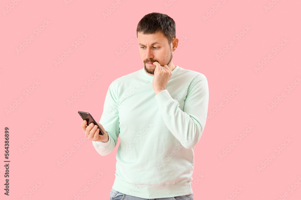 Handsome man with mobile phone biting nails on pink background