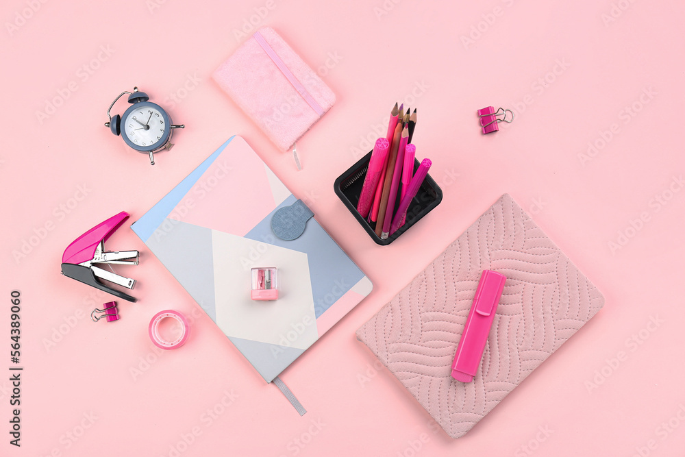 Holder with stationery, notebooks and alarm clock on pink background