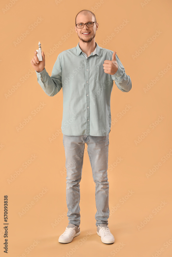 Young man in eyeglasses with disposable electronic cigarette showing thumb-up on beige background