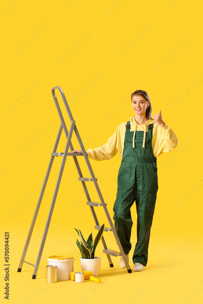 Young woman with ladder, houseplant and paint cans showing thumb-up on yellow background