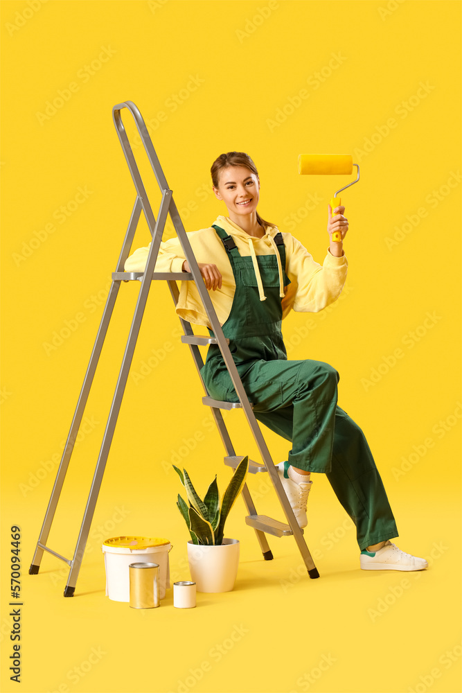 Young woman with roller, ladder, houseplant and paint cans on yellow background