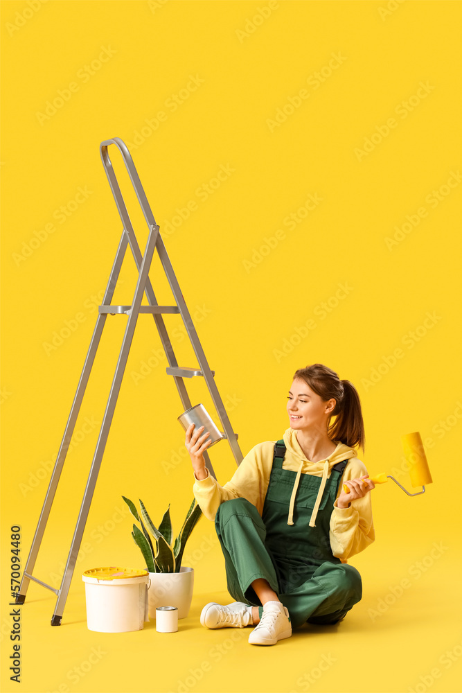 Young woman with roller, ladder, houseplant and paint cans on yellow background