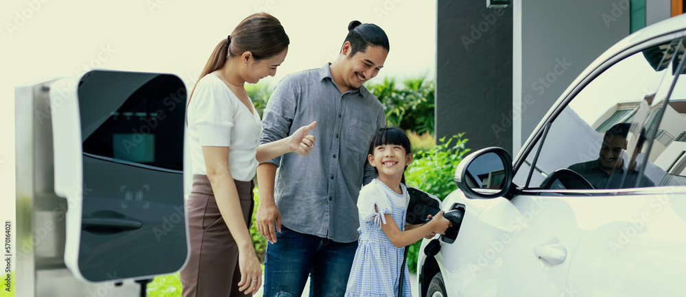 Progressive young parent teach daughter how to recharge or refuel EV car at home charging station. G