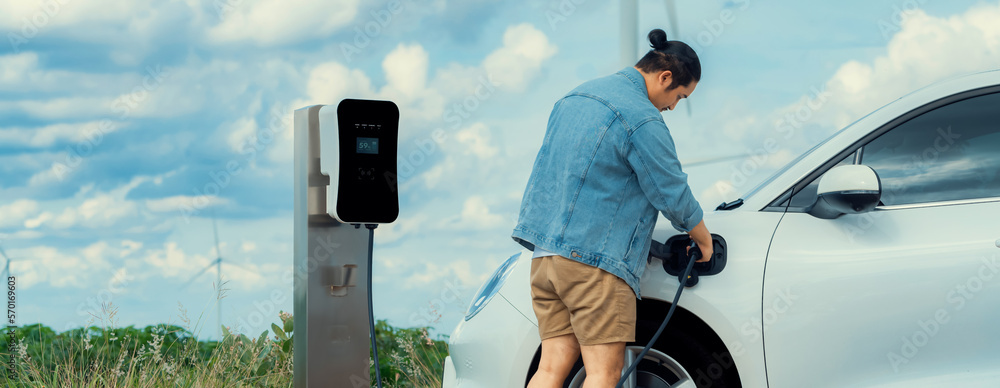 Progressive man with his electric car, EV car recharging energy from charging station on green field