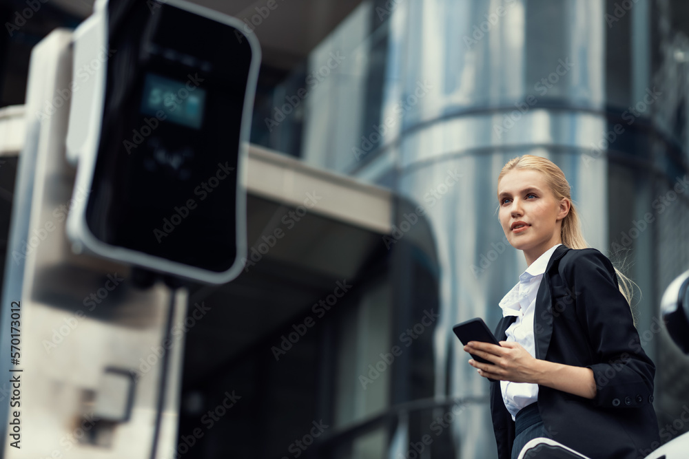 Businesswoman wearing black suit using smartphone, leaning on electric car recharge battery at charg