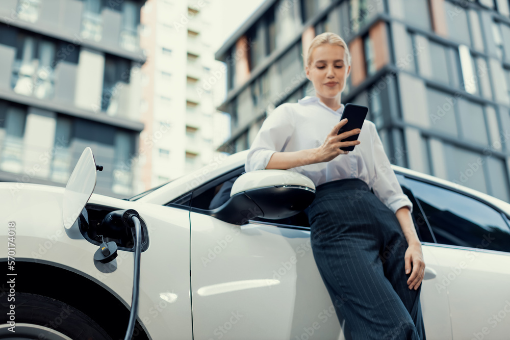 Closeup progressive businesswoman talking on smartphone, leaning electric car at charging station wi