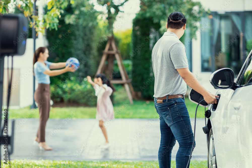 Focus image of progressive man charging electric car from home charging station with blur mother and