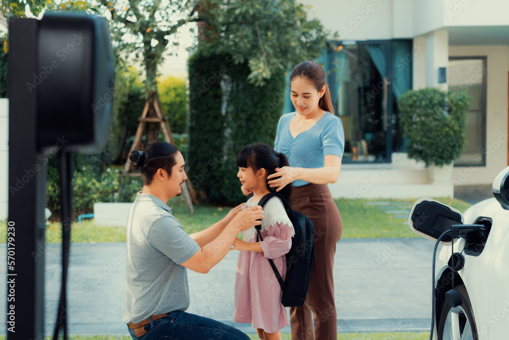 Progressive young parents and daughter with electric vehicle and home charging station. Green and cl