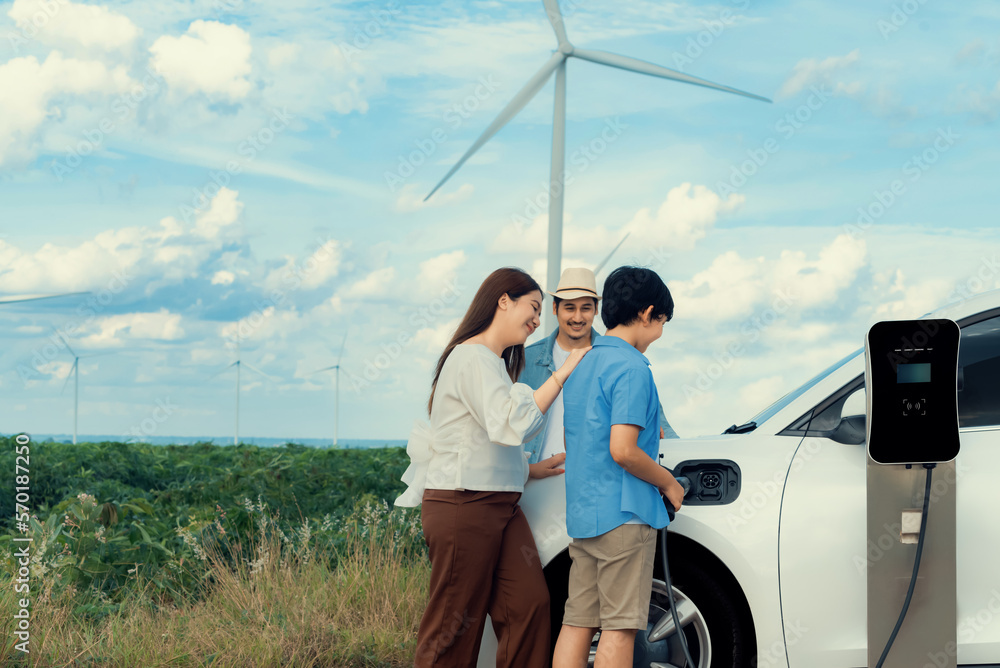 Concept of progressive happy family enjoying their time at wind farm with electric vehicle. Electric