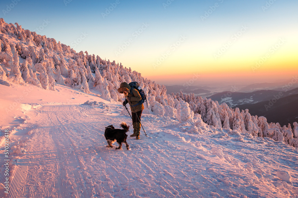 Woman with her dog hiking in winter mountain during sunset. Trekking in snowy landscape. Sport and a