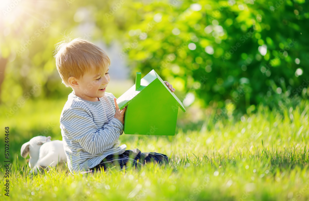 Smiling child sitting on the spring meadow with model of the green house in his hands.