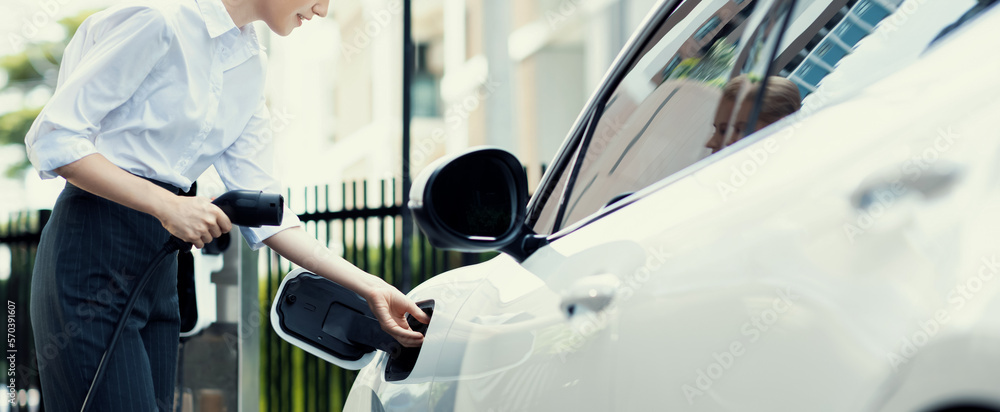 Closeup progressive businesswoman with smartphone, leaning electric car and charging station before 