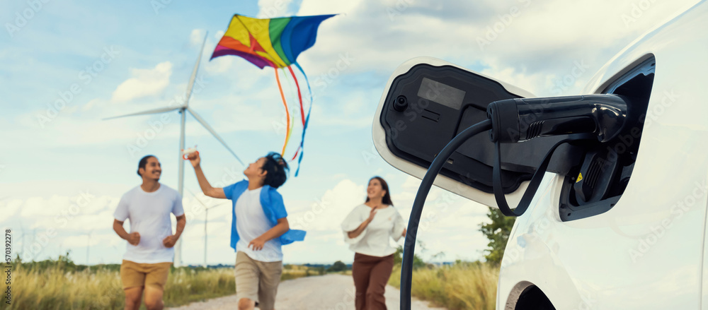 Concept of progressive happy family holding windmill toy and relax at wind farm with electric vehicl