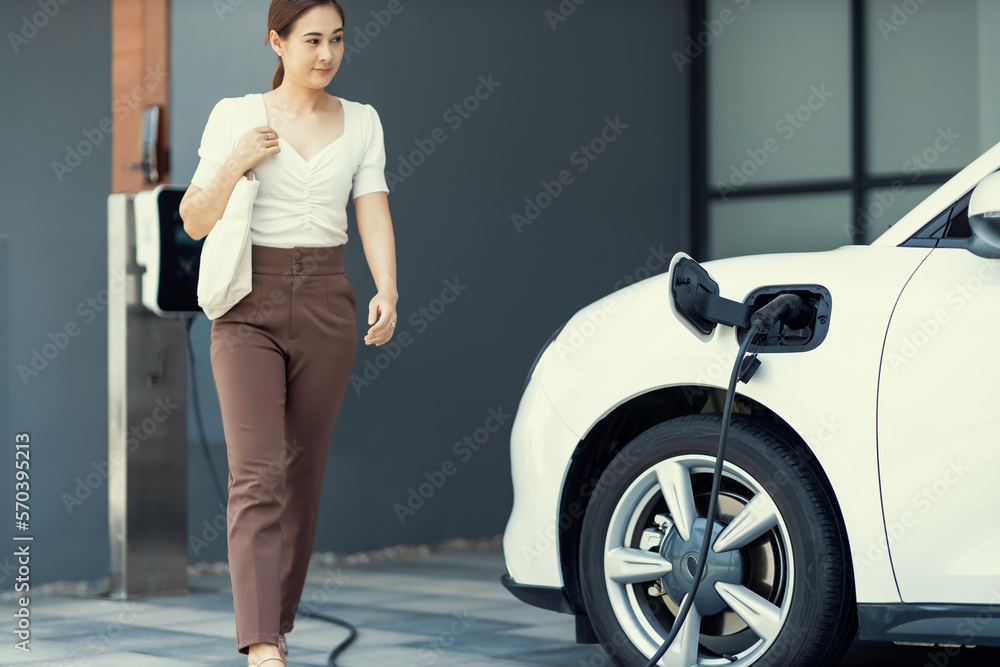 Focus image of electric vehicle recharging battery at home charging station with blurred woman walki