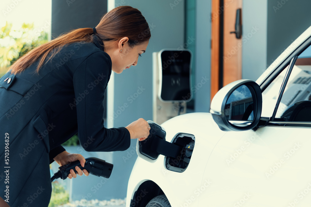 Progressive woman install cable plug to her electric car with home charging station. Concept of the 