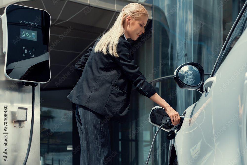 Progressive businesswoman insert charger plug from charging station to her electric vehicle with apa